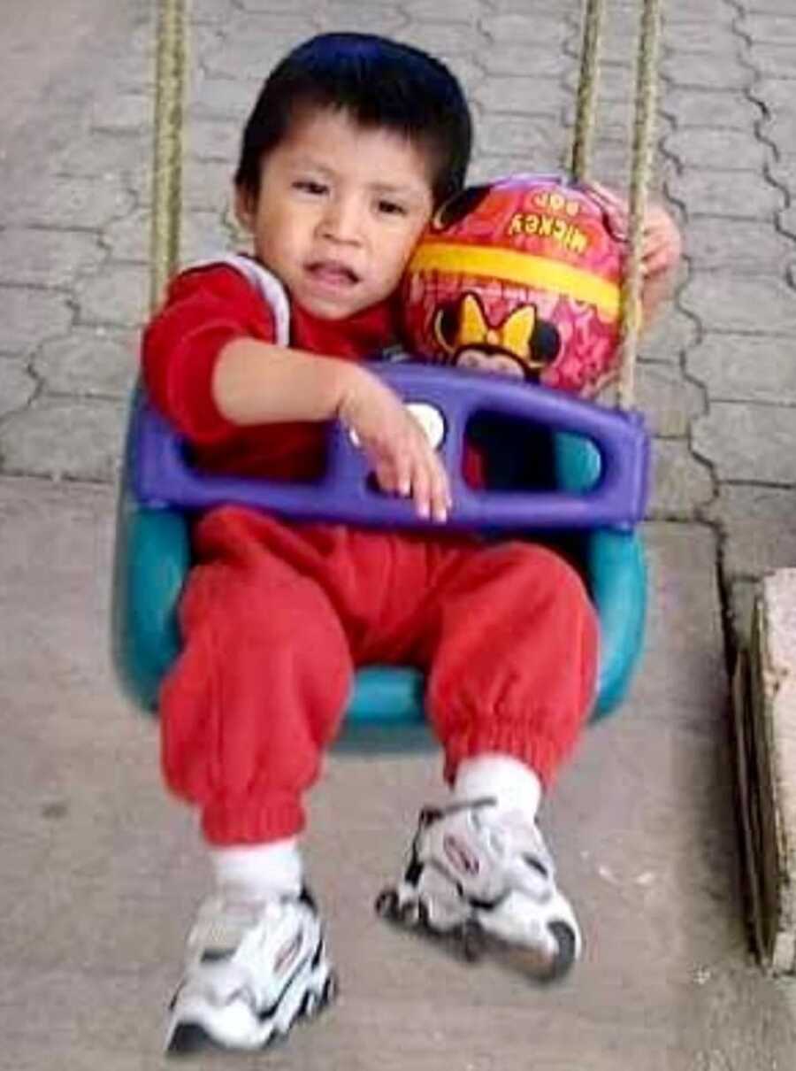 Baby boy sits in plastic swing and holds ball next to his face.