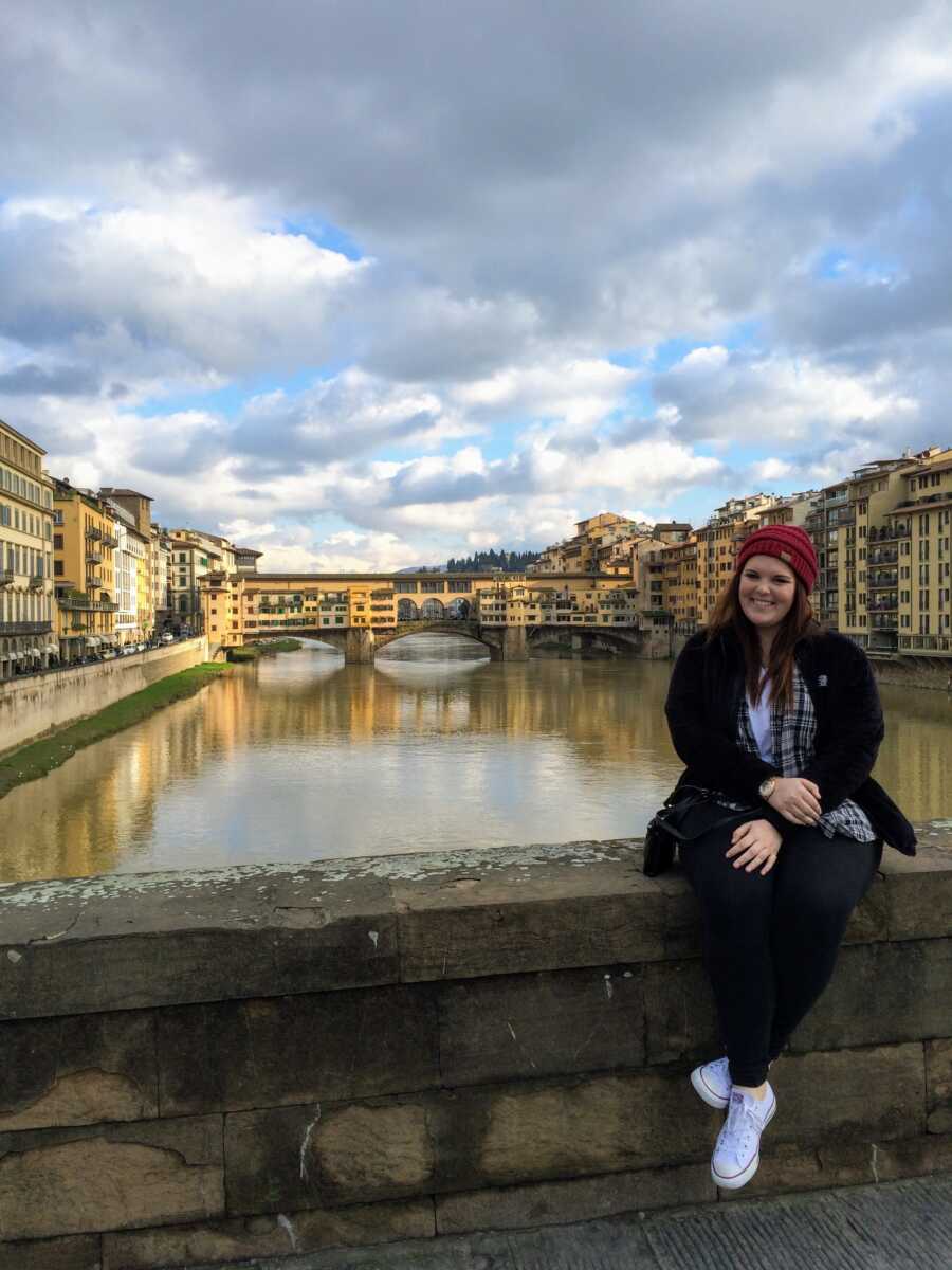 Young woman sits on a bridge in front of a river and old buildings