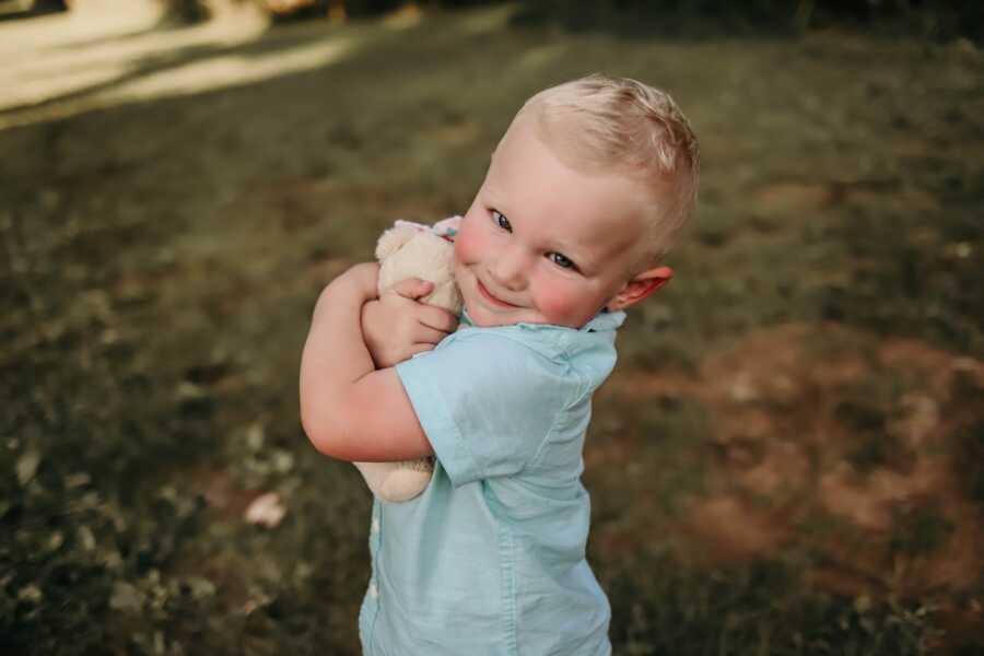 young boy stands holding tight onto a teddybear with a soft smile