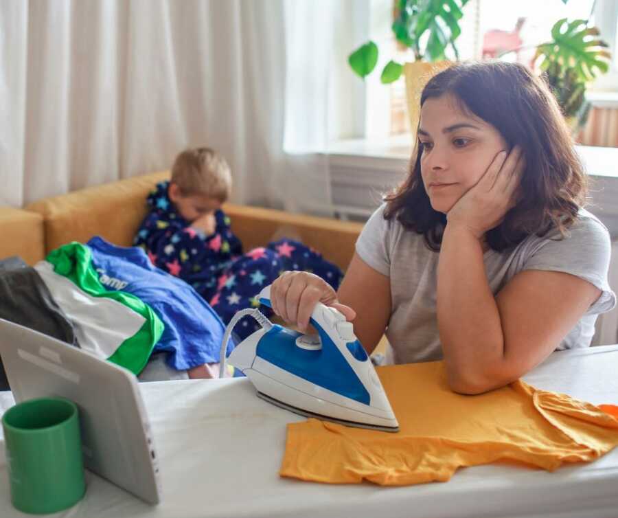 Mom irons clothes while watching video on tablet while son sits behind her on the couch.
