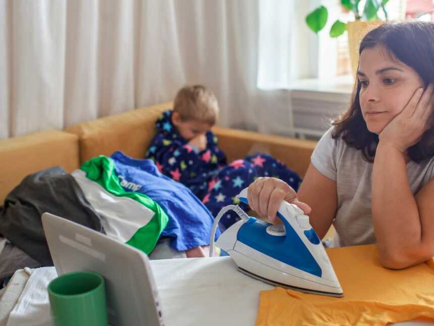 Mom irons clothes while watching video on tablet while son sits behind her on the couch.