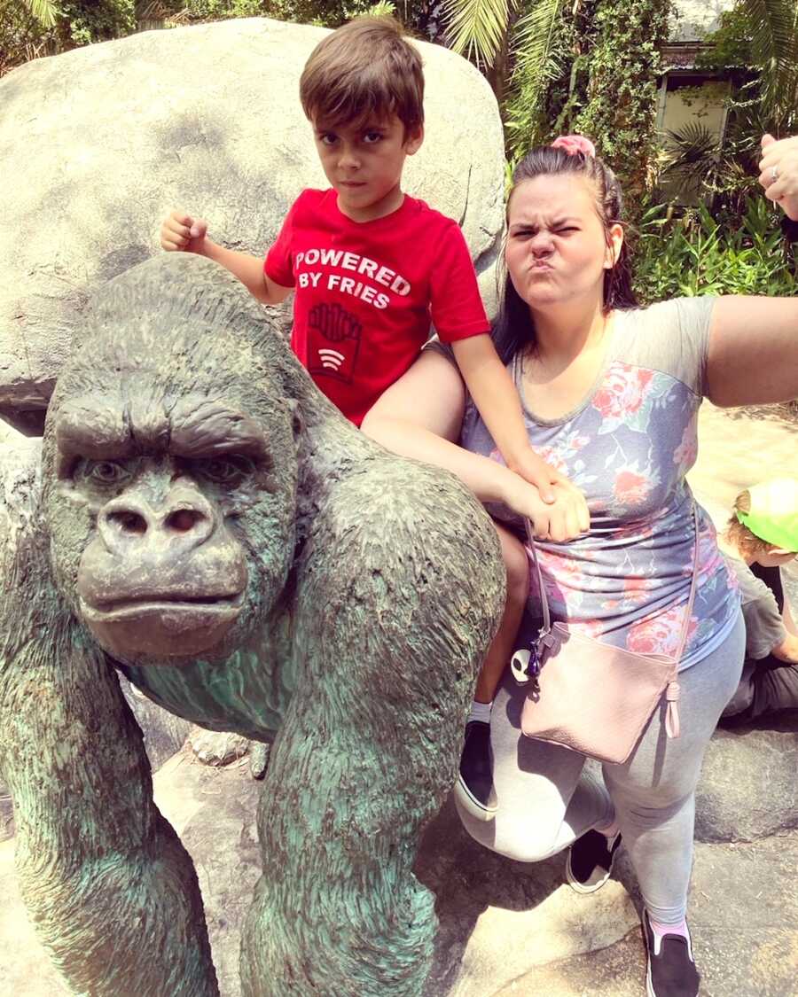 son sits on gorilla statue making serious face while mom makes a tough face