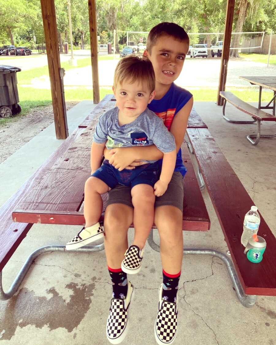 older brother holds his little brother while sitting on picnic table