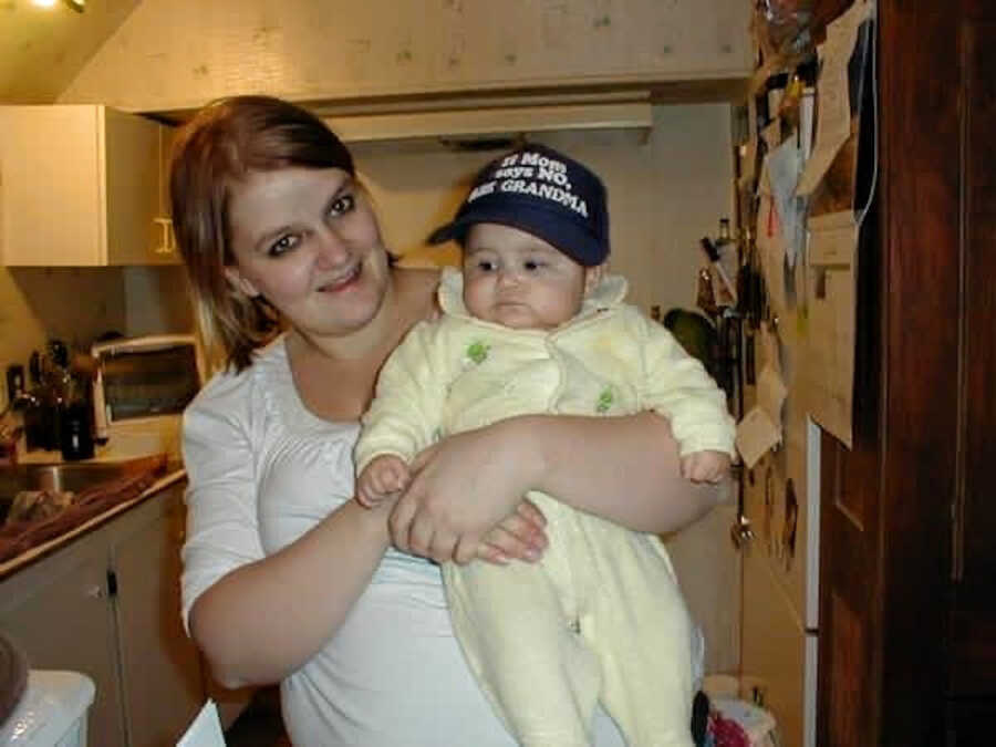 A young woman holds a baby dressed in yellow and a ballcap