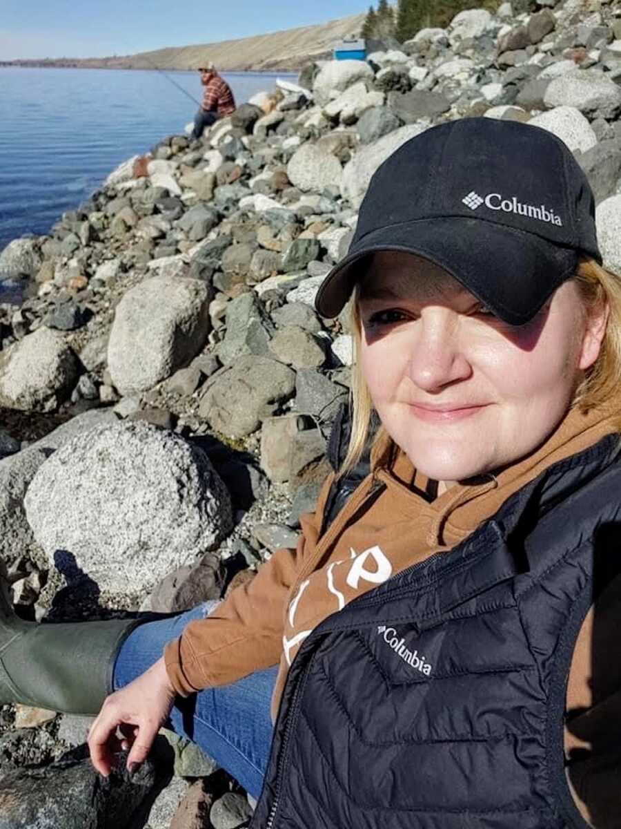 A woman sits on a rocky shore smiling at the camera