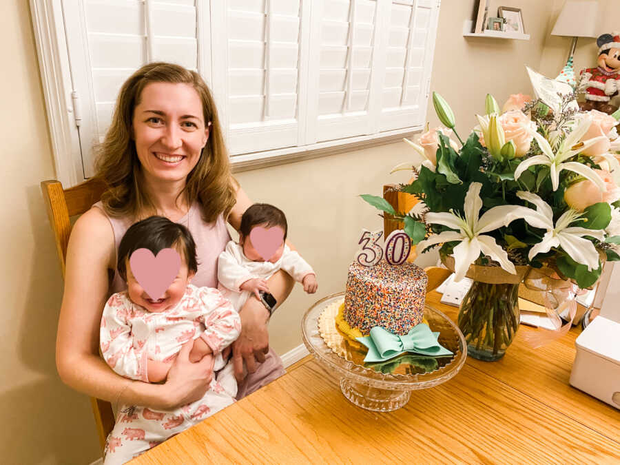 Single foster mom holds baby sisters as she sits at table with cake and flowers for her 30th birthday.