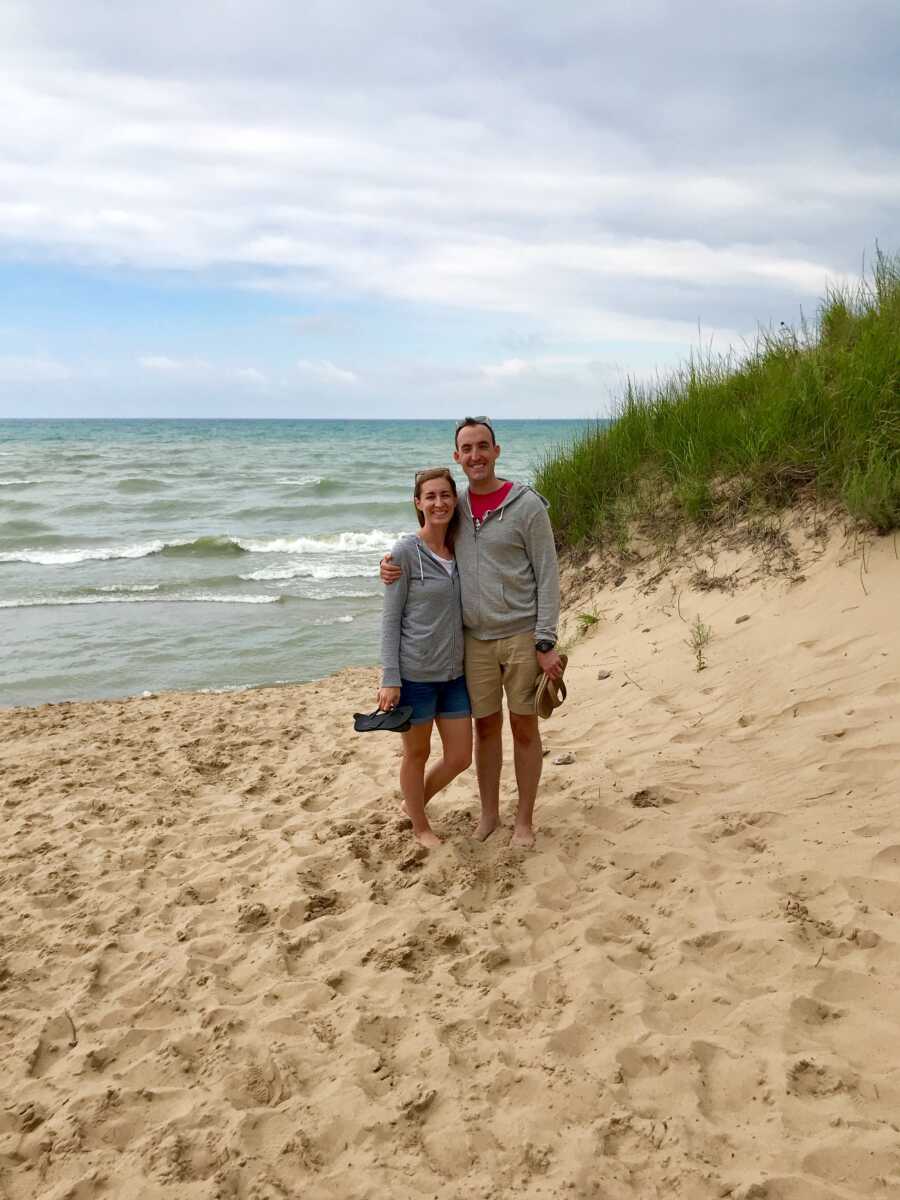 Couple stand together at the beach, holding their shoes as they stand in the sand.