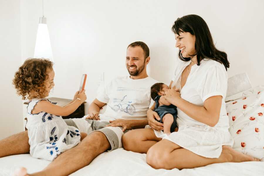 family of four sits in bed together, mom holds baby while father entertains young child