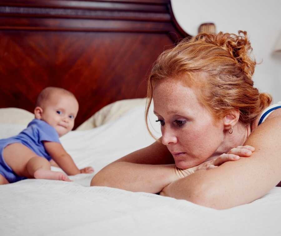 Young mom lies on bed resting her head on her arms while a young baby crawls around behind her.