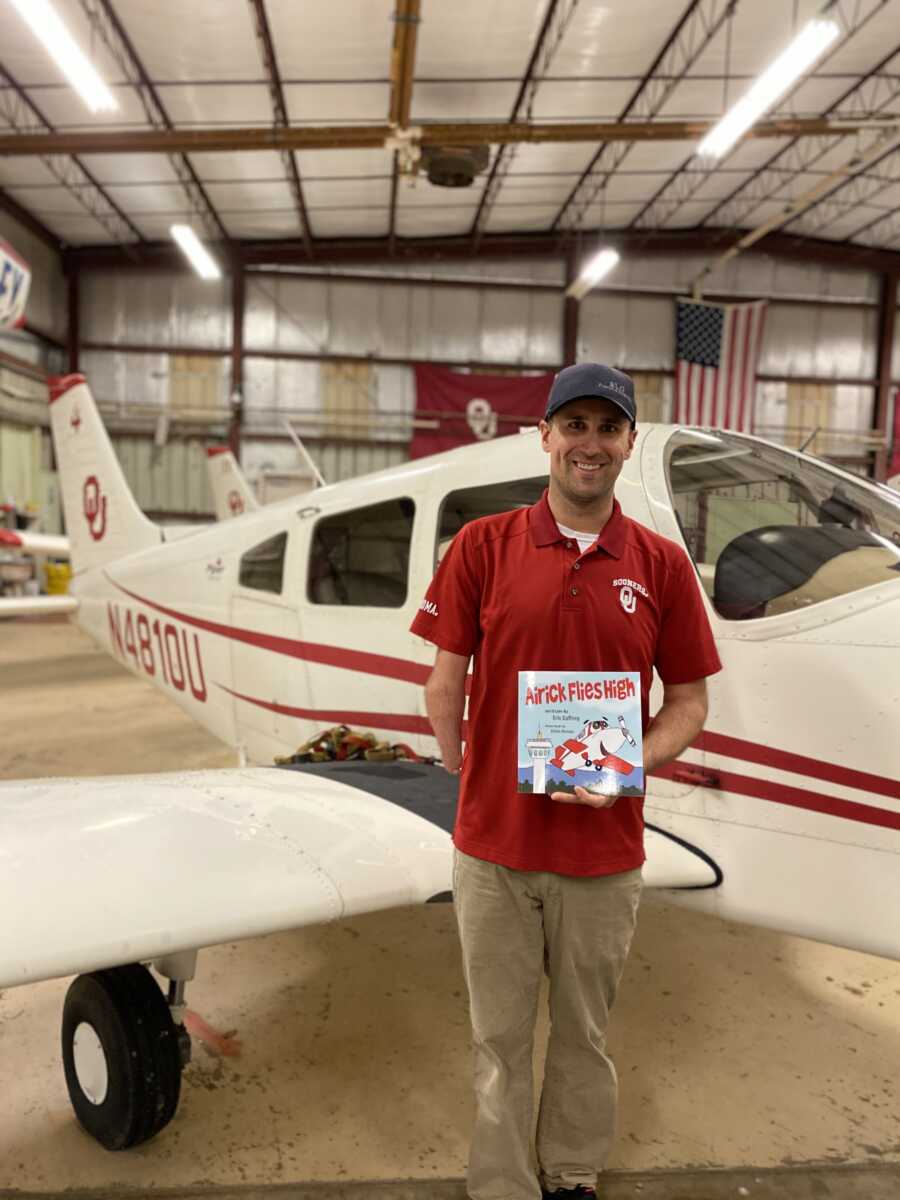 Pilot with limb difference stands in front of plane holding up children's book he wrote about his journey.