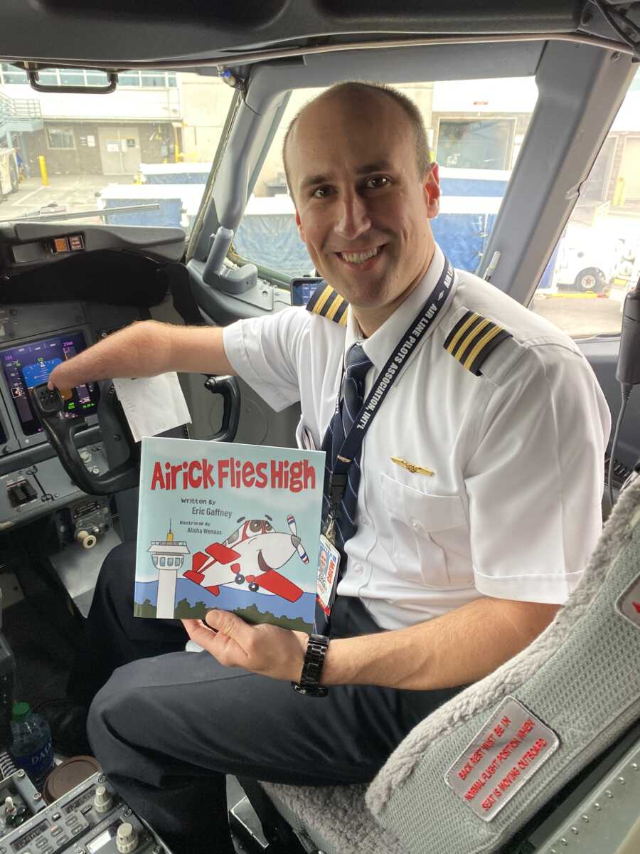 Pilot sits in cockpit holding children's book he wrote about cartoon airplane with limb difference.