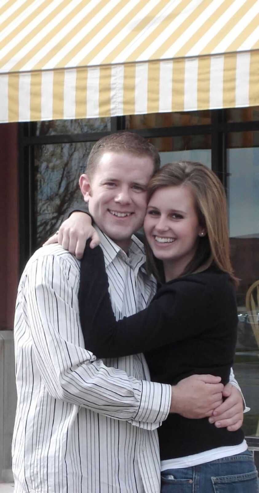 Young wife wraps arms around husband's neck and takes picture under awning.