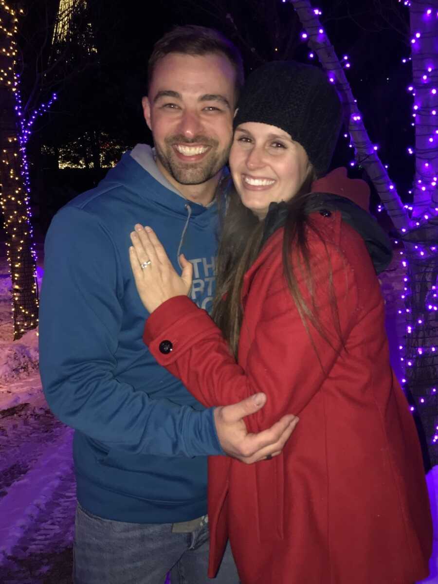 Newly engaged couple take picture holding each other next to trees with Christmas lights.
