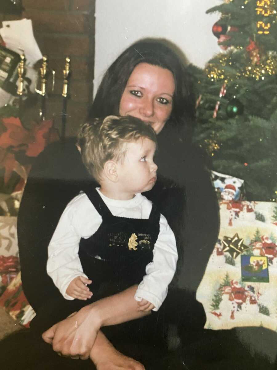 Young mom holds toddler boy wearing black overalls on her lap while sitting in front of Christmas tree and presents.