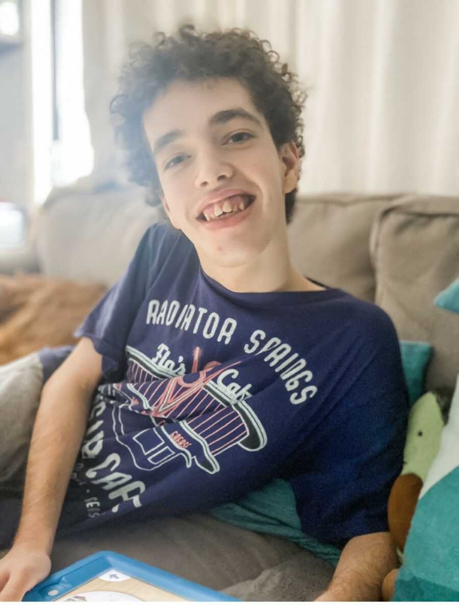 Nonverbal teenager with rare genetic disease sits on couch with ipad and smiles at camera.