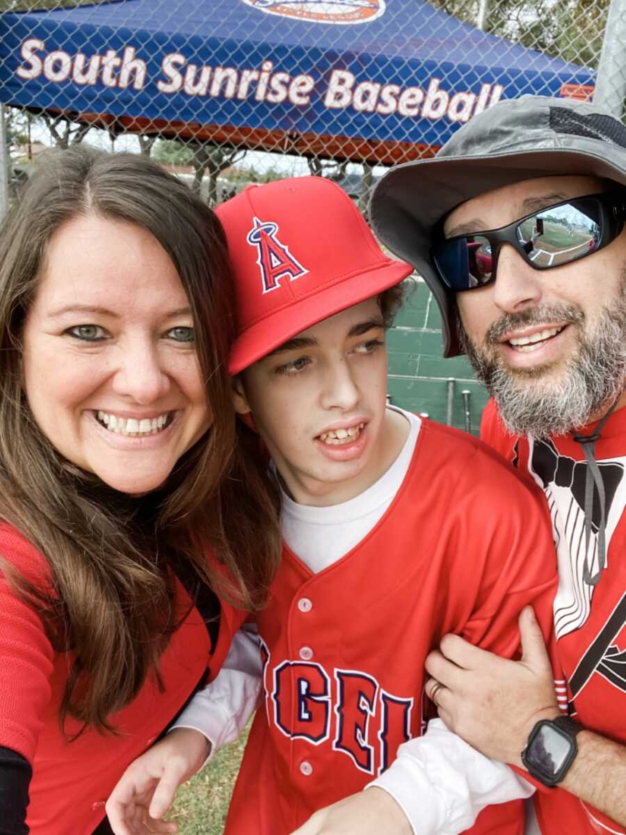 Parents take picture on either side of teen with special needs at a baseball game.