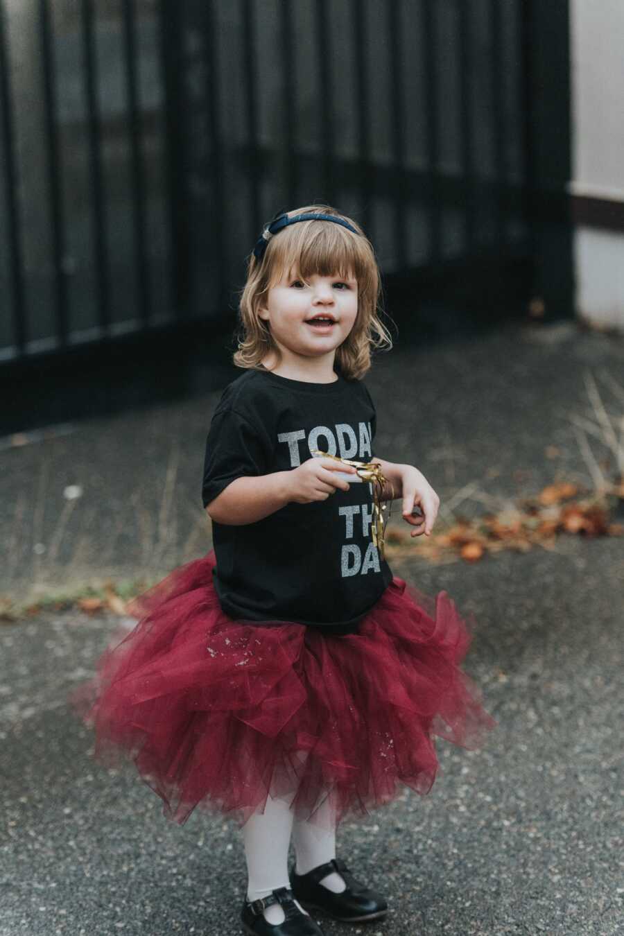 An adopted little girl in a tutu stands on pavement in front of black gate