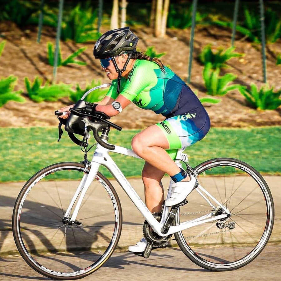 Woman in green and blue body suit rides bike with one arm