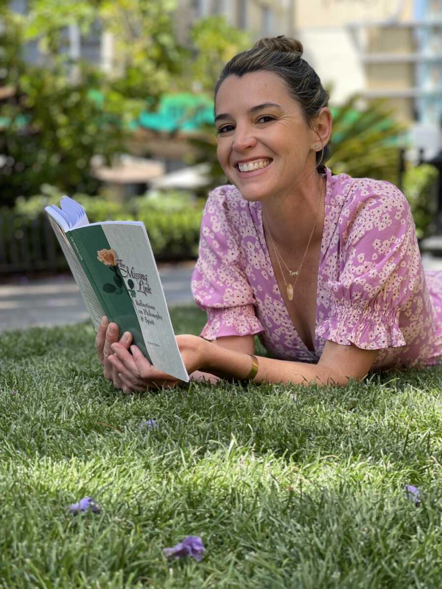 A woman in a pink dress lays in the grass reading and smiling