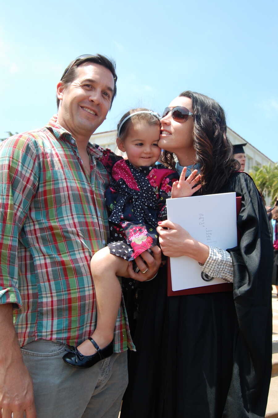 A woman in graduation robes holds her daughter with her husband