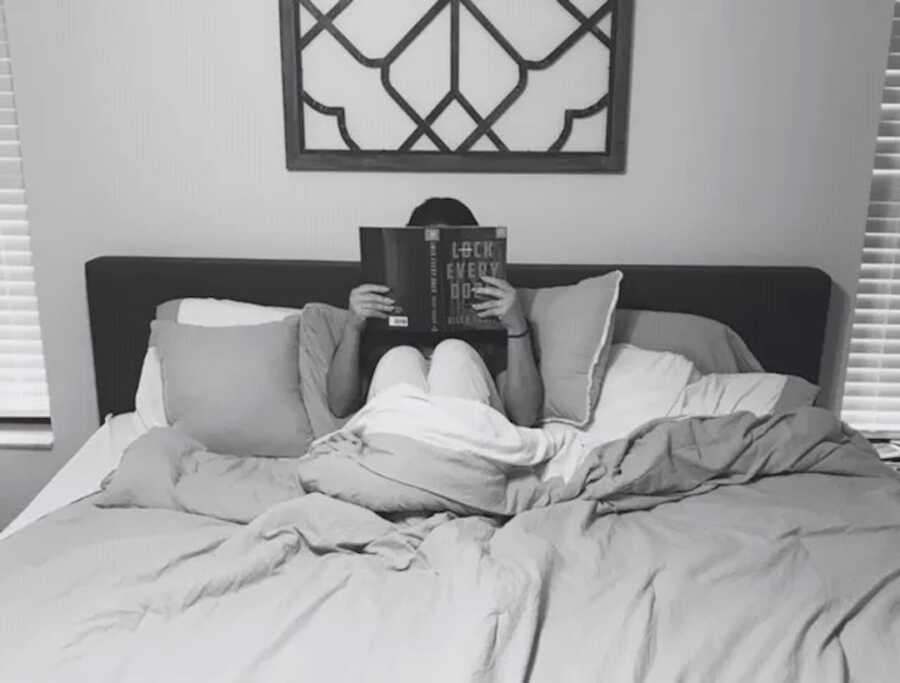 woman sits in bed under covers with a book covering her face