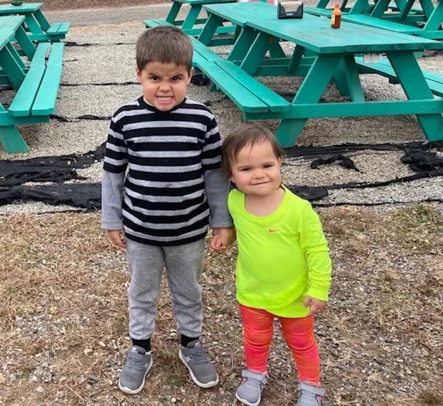 siblings stand by picnic tables holding hands, daughter smiles while son makes a silly face