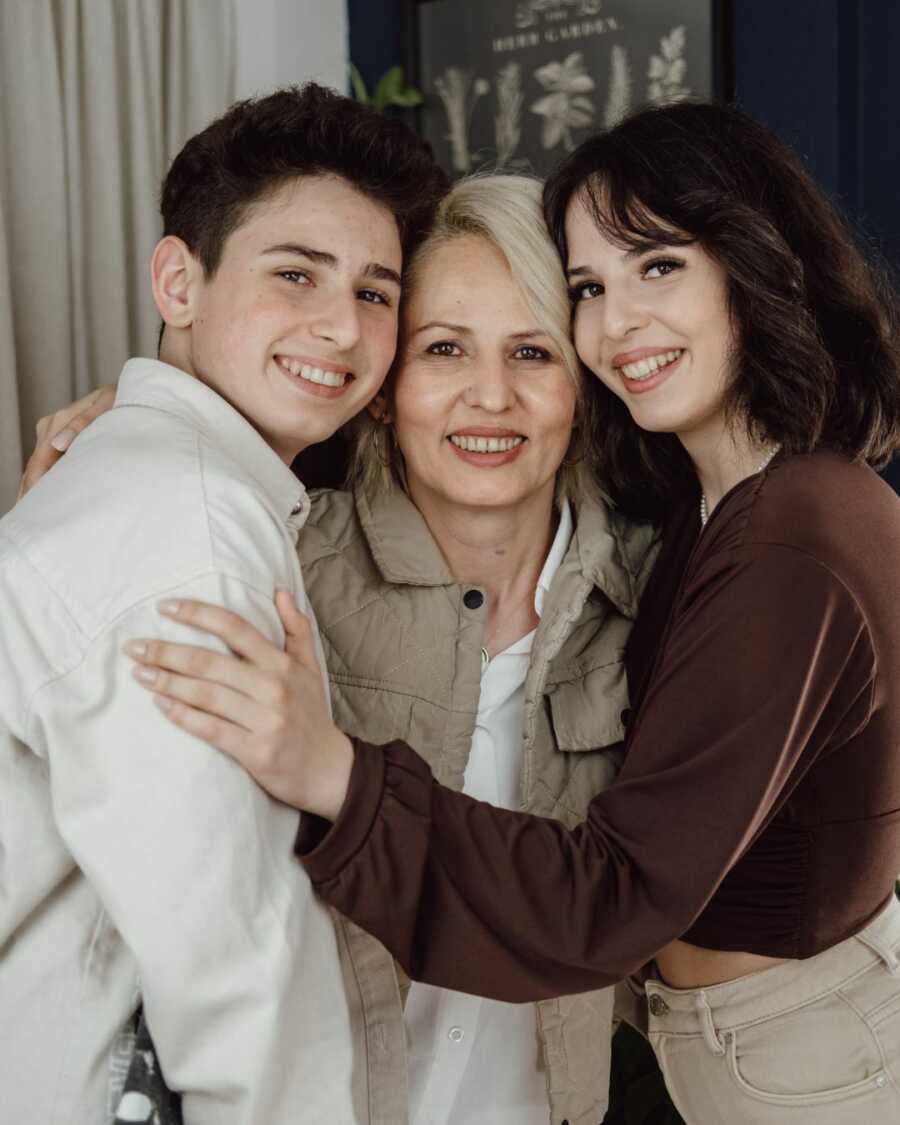 Middle-aged mom receives hugs from teen son and daughter on either side of her.