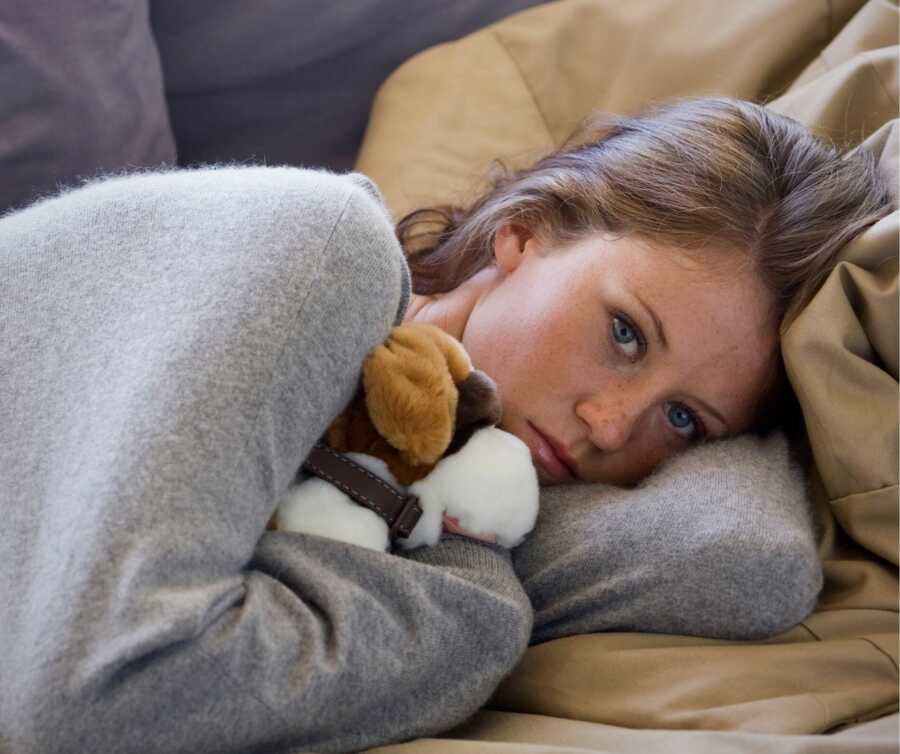 Depressed teenage girl lies on couch with dog stuffed animal pulled tightly to her chest.