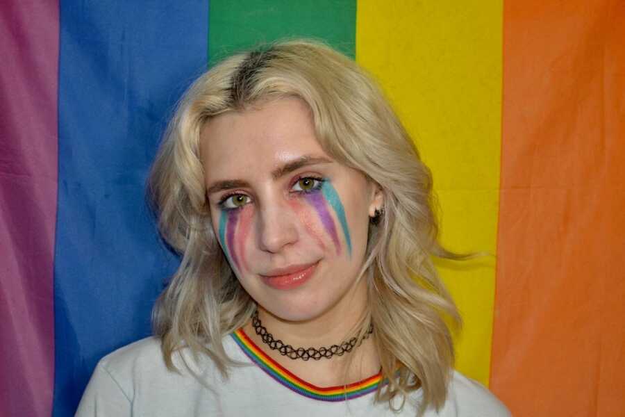 Mental health advocate stands in front of a pride flag with the bisexual flag colors facepainted on