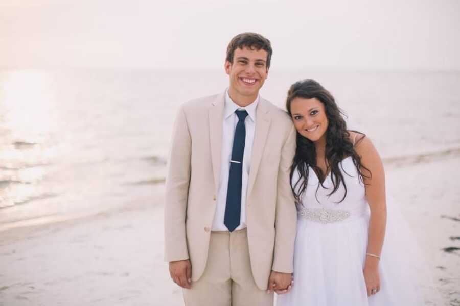 Bride and groom lean into each other and hold hands on the beach.