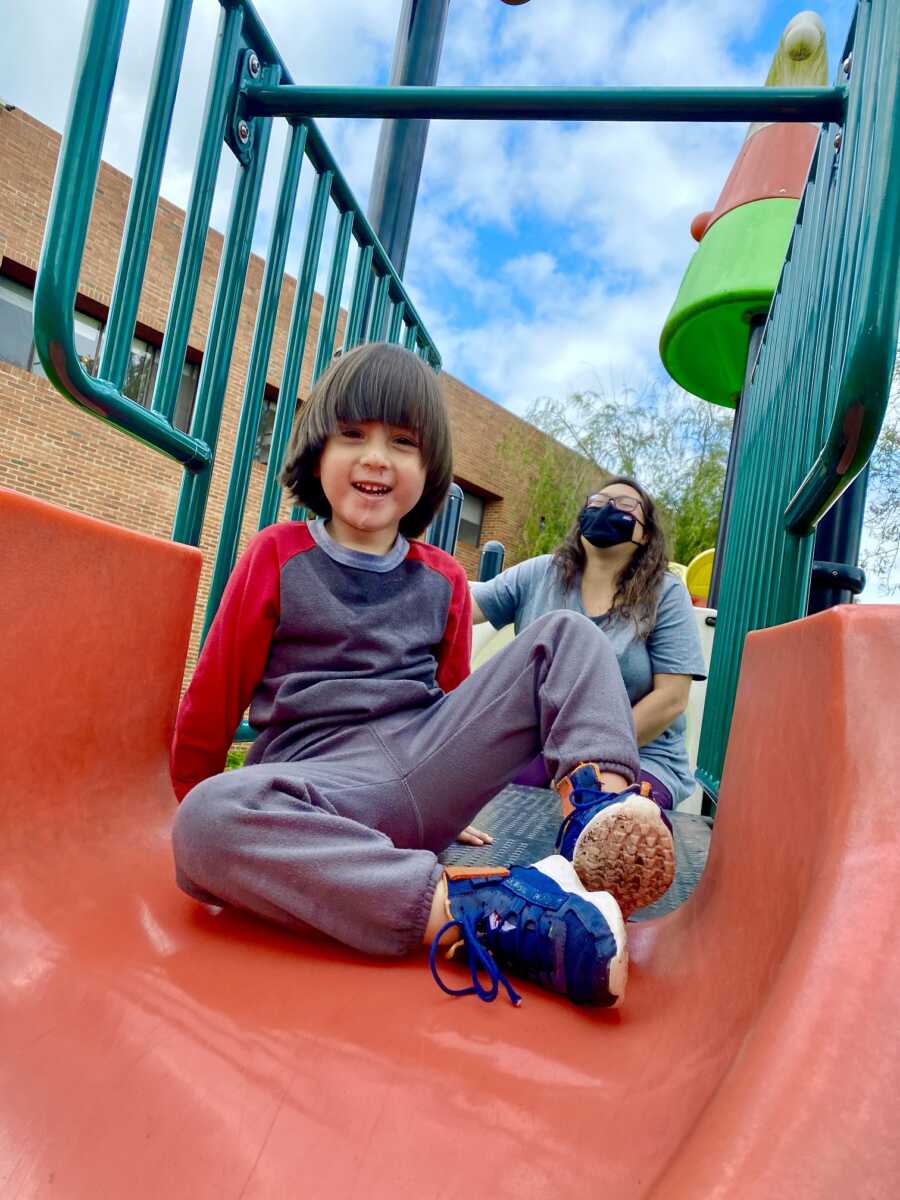 Young boy goes down slide at orphanage with adoptive mom standing behind him.