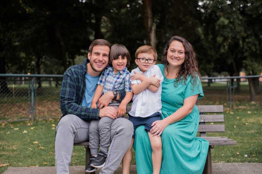 Adoptive parents take picture with two young boys sitting on a park bench.