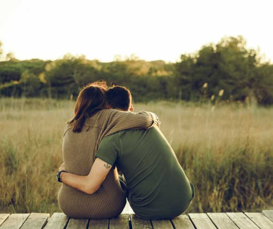 Husband and wife sit on dock and hug each other while looking out at the trees and grass field.