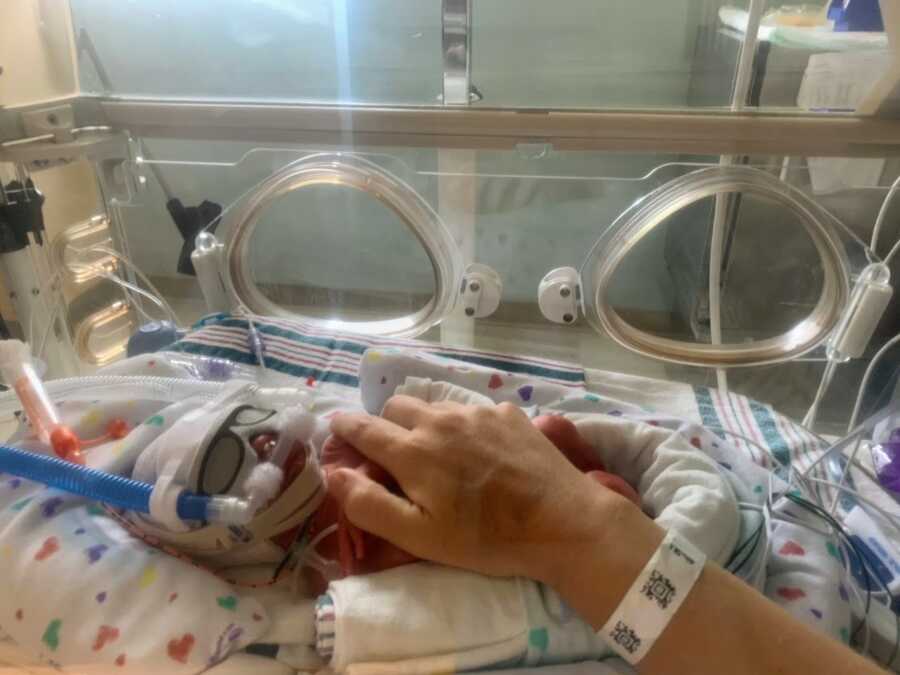A mom's hand holds over her baby hooked up to oxygen in the NICU