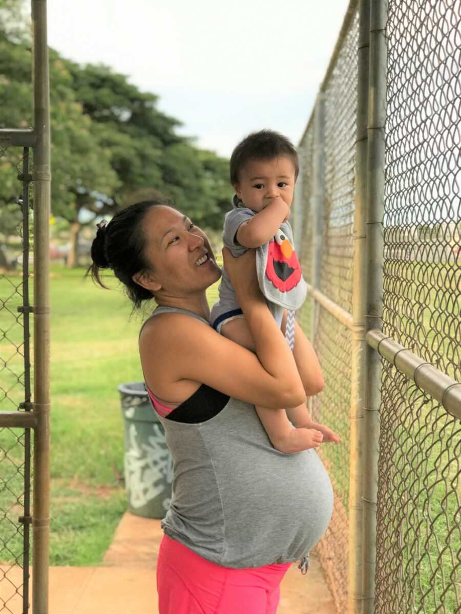 Pregnant woman holds up young baby boy at the park.