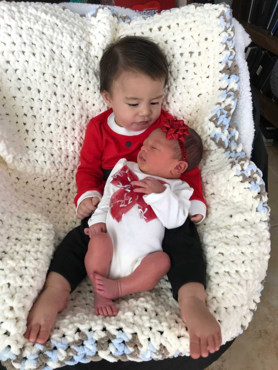 Baby boy holds newborn baby sister in his lap with both wearing cute matching red outfits.