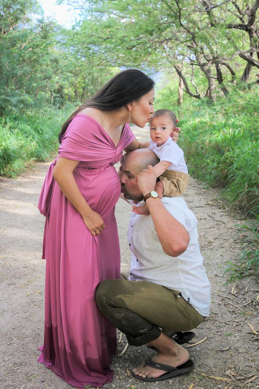 Husband kisses wife's pregnant belly while holding baby boy on top of his shoulders.