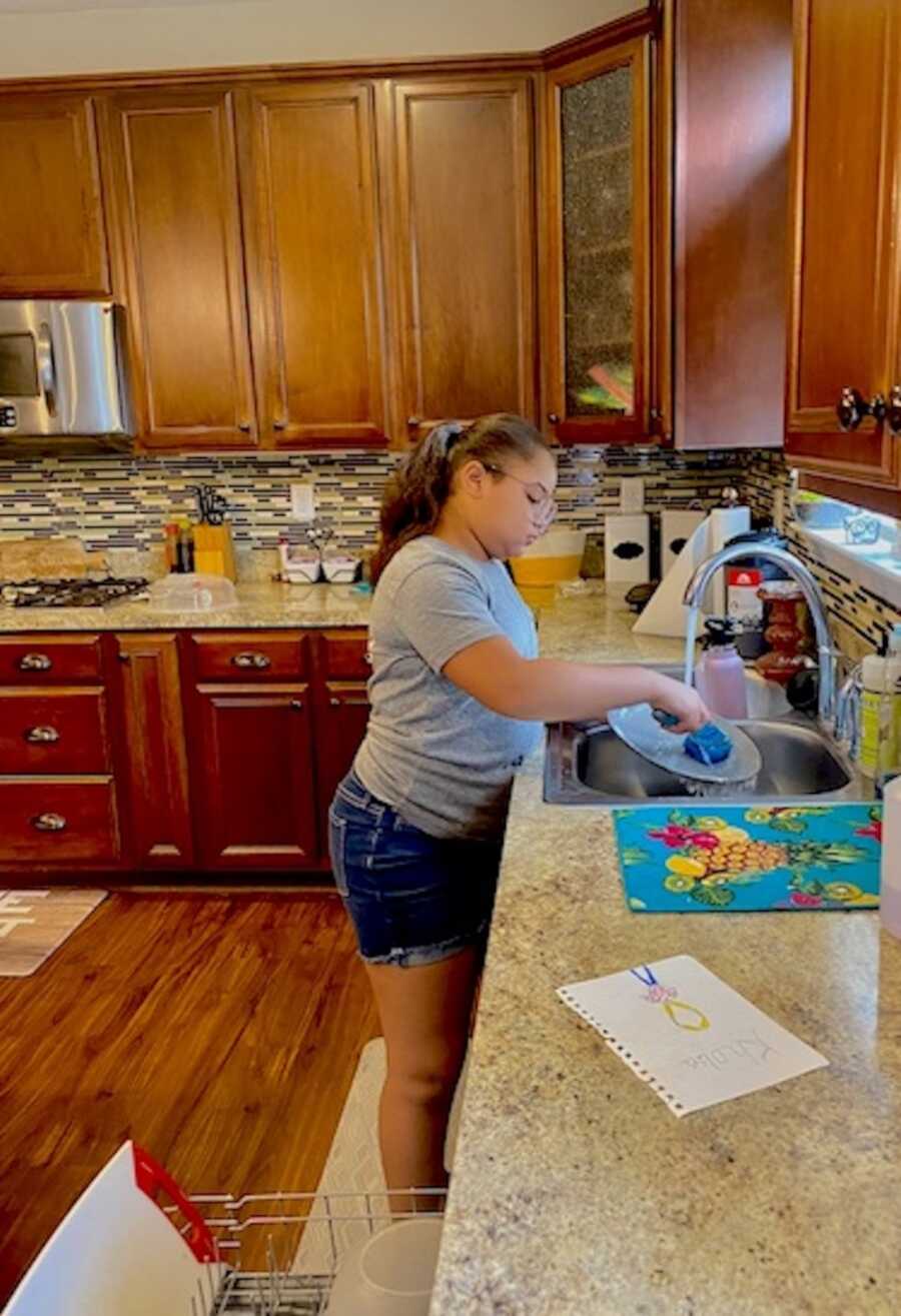 young girl stands at sink washing dishes, completing chores