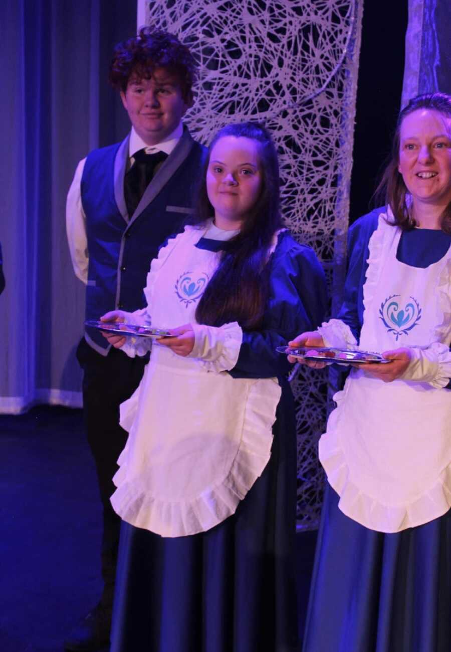Student actors in Frozen performance stand holding trays as castle staff.