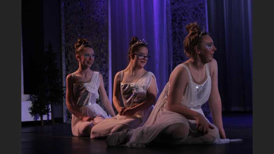 Three girls in costume sit on stage watching other actors.