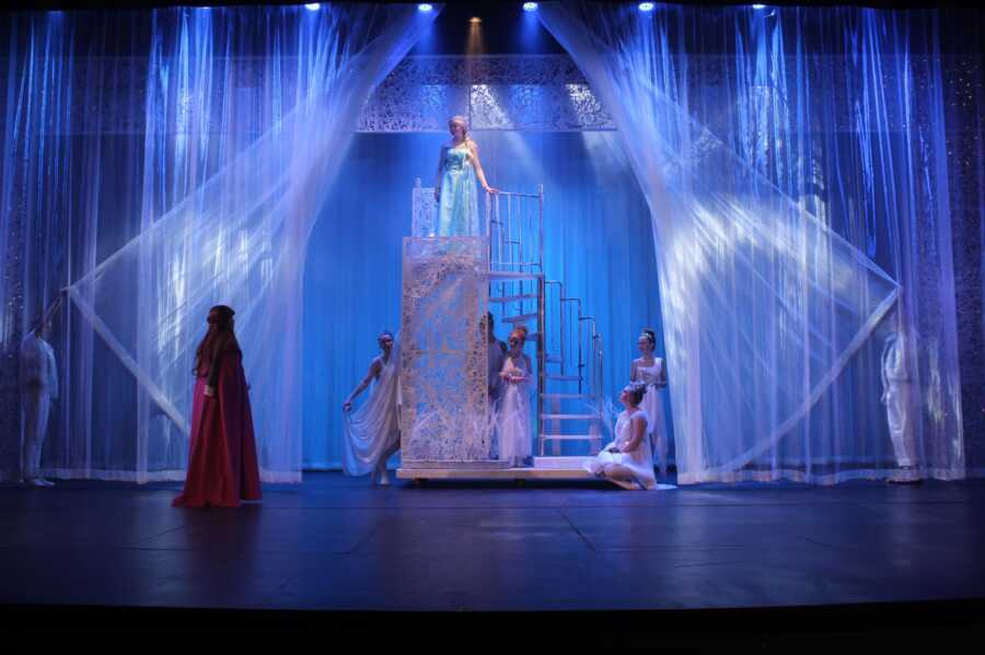 Student actor playing Elsa from Disney's Frozen stands on an ice block prop on stage.
