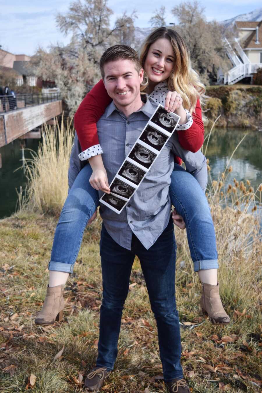 husband giving wife a piggyback ride while she holds ultrasound image