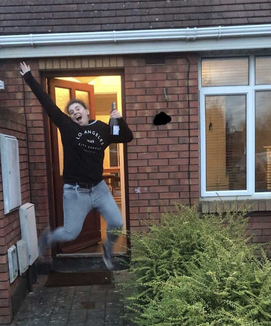 Woman jumps in doorway of house, excitedly holding a bottle of wine.