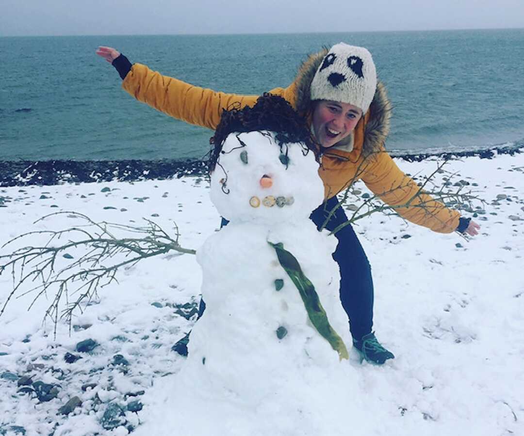 Young woman stands behind a snowman with her arms spread out.