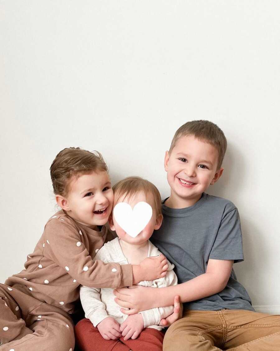 Siblings wrap arms around foster baby while sitting on the floor with their backs against a white wall.