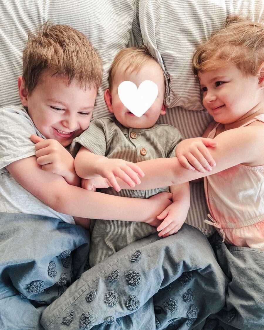 Siblings wrap arms around baby foster brother while snuggling in bed.
