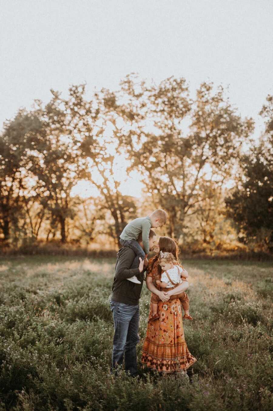 Couple kiss while wife holds daughter in her arms and husband holds son on his shoulders.