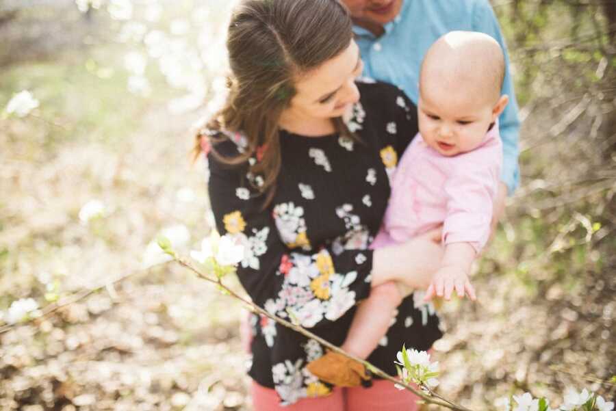 Mom holds baby girl reaching for blossom on tree branch.