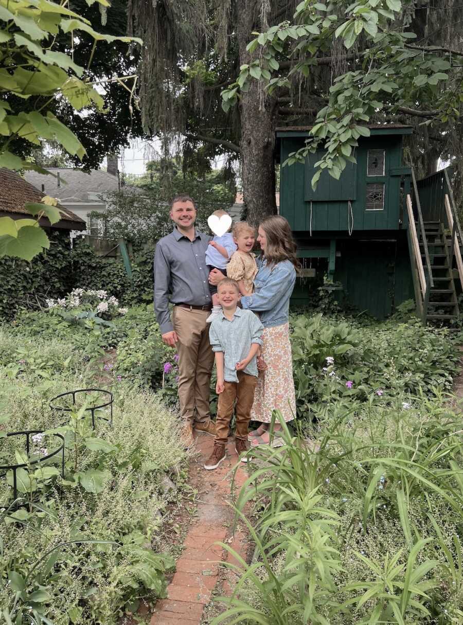 Family stands in outdoor garden and shrubbery with dad holding foster son.