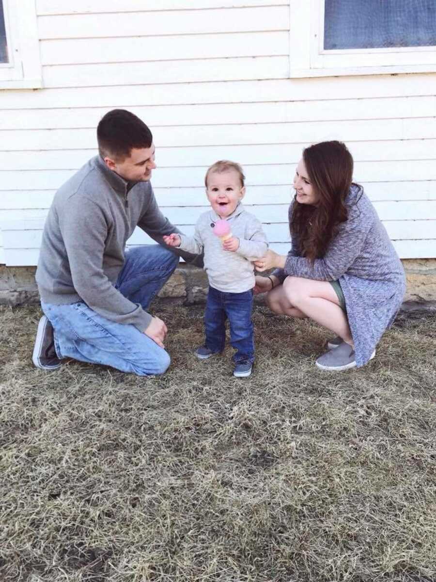 Parents crouch down in the grass next to toddler son.